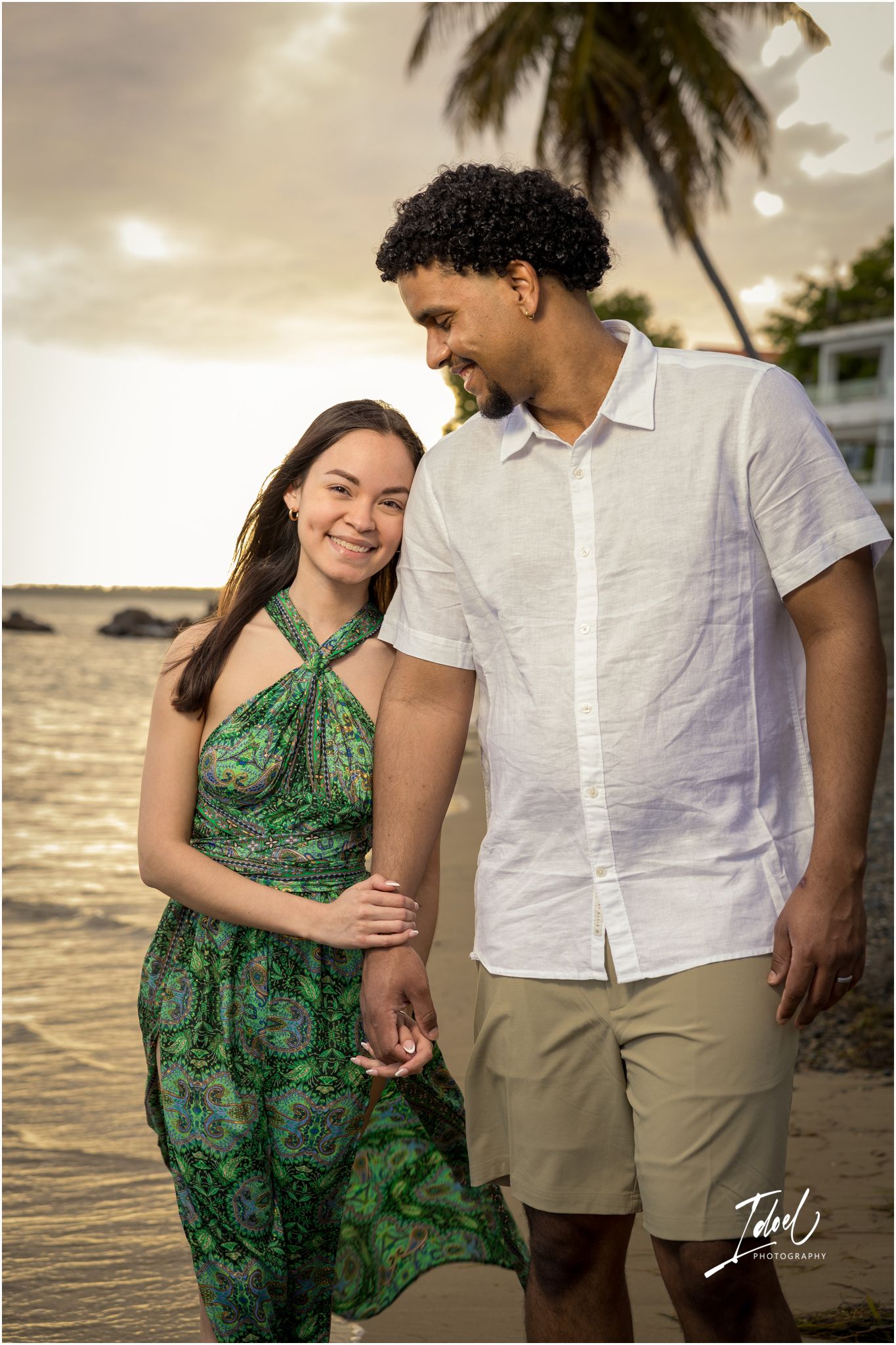 A couple walks together on a tropical beach at sunset with a palm tree in the background.