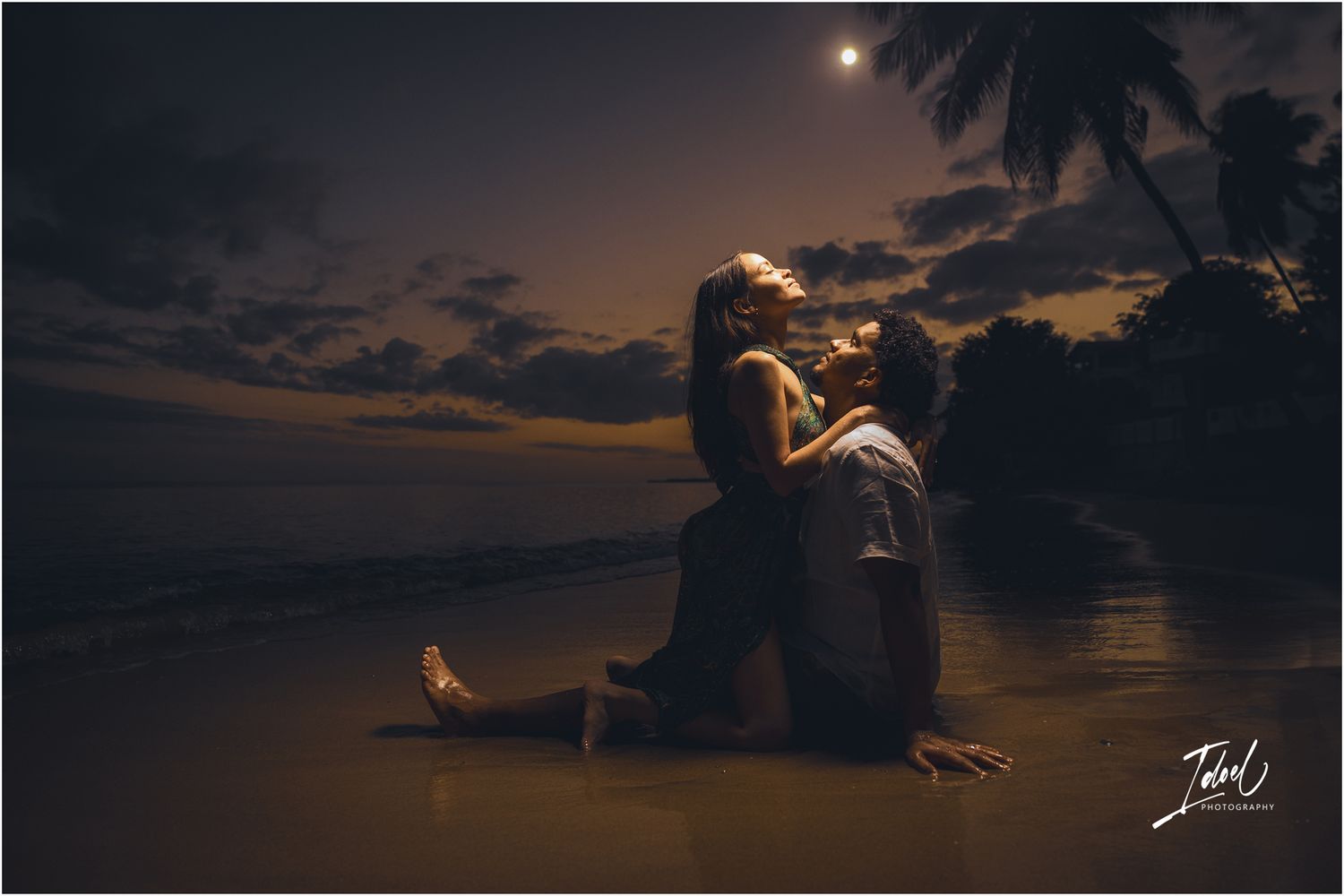 A silhouetted figure sits on a beach at night with a glowing moon and palm trees in the tropical background.