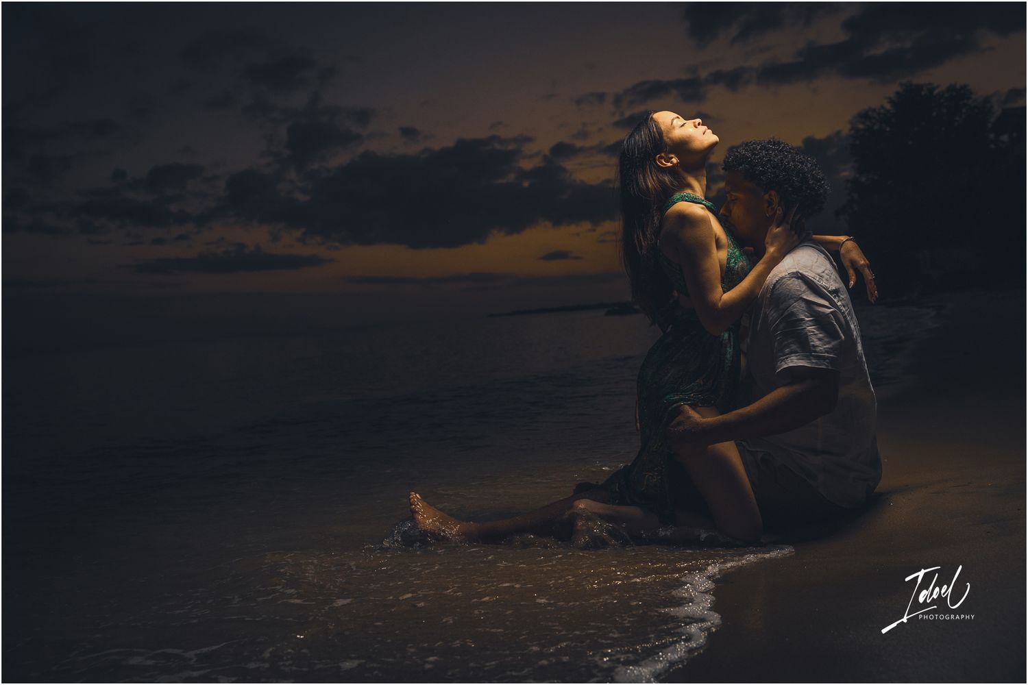 Silhouette of a couple sitting by the ocean shore at sunset with dramatic dark clouds overhead.