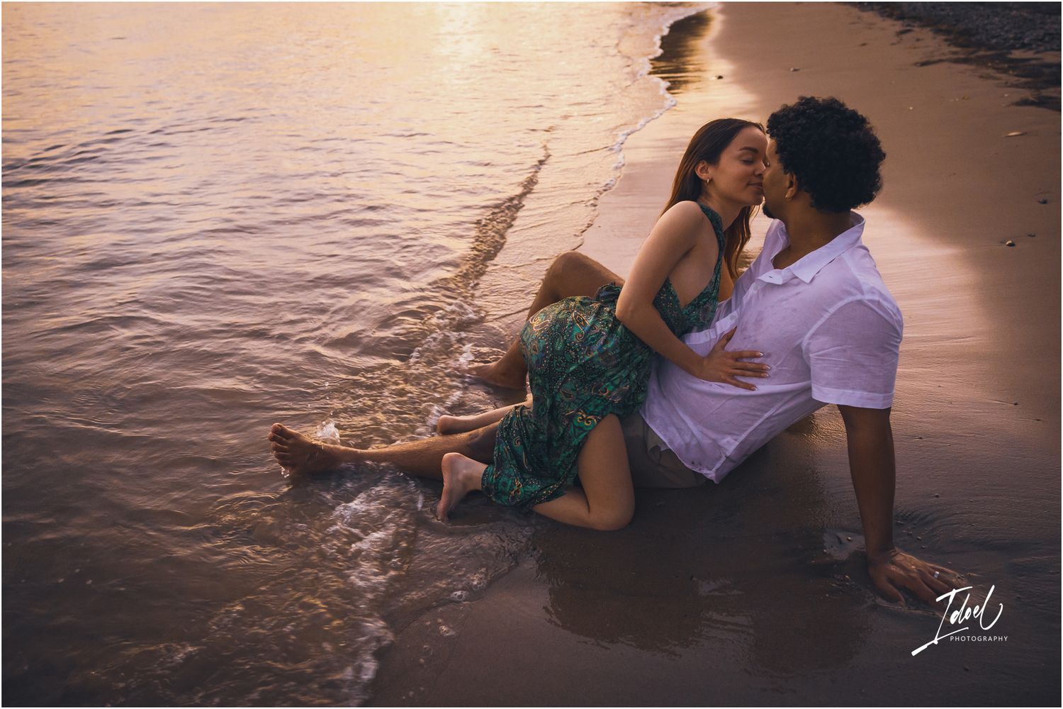 A romantic couple sits in shallow ocean water at sunset, waves gently lapping around them on the beach.