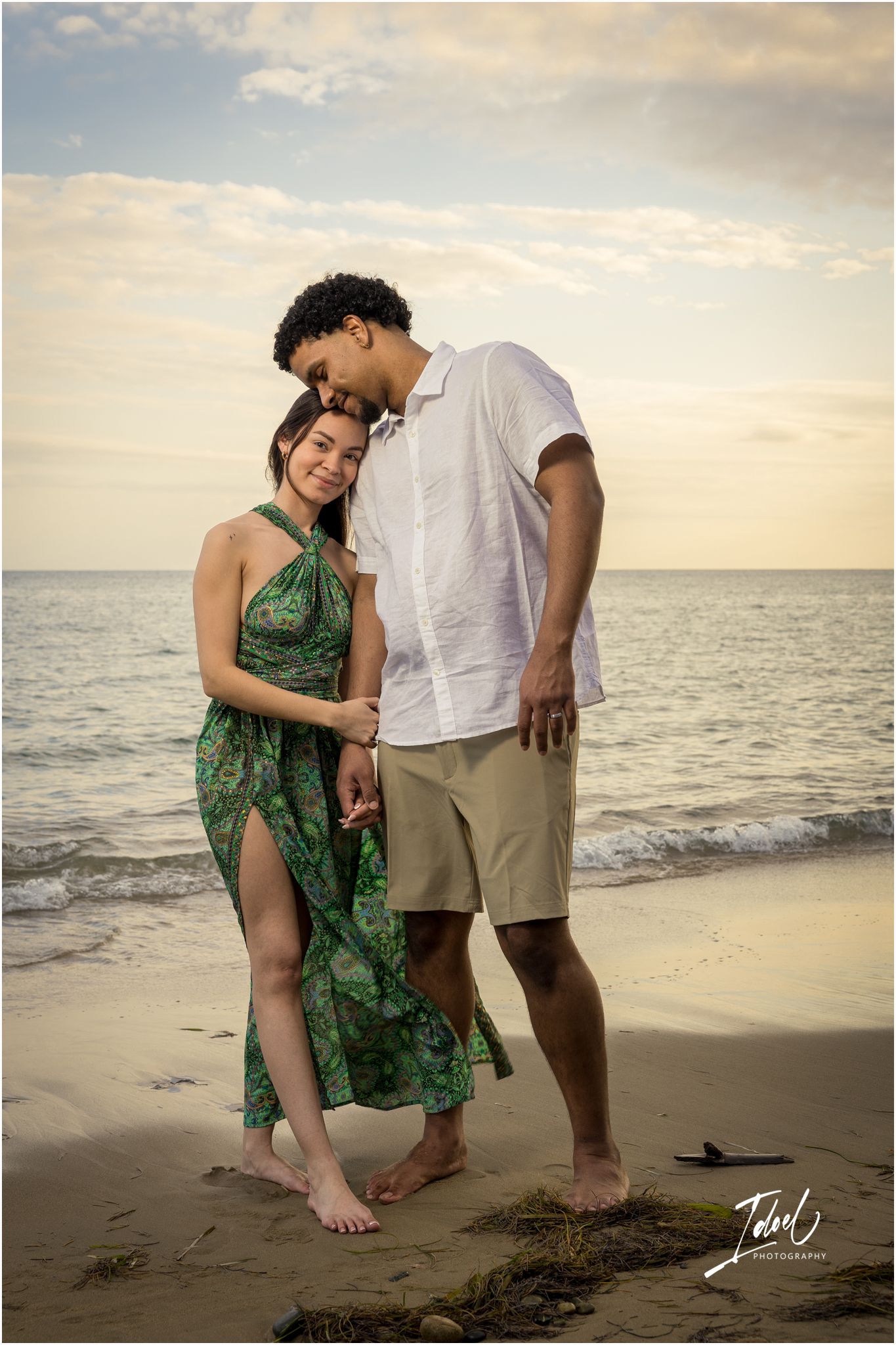 A couple shares an intimate moment on a beach at sunset, with the woman wearing a flowing green dress and man in casual attire.