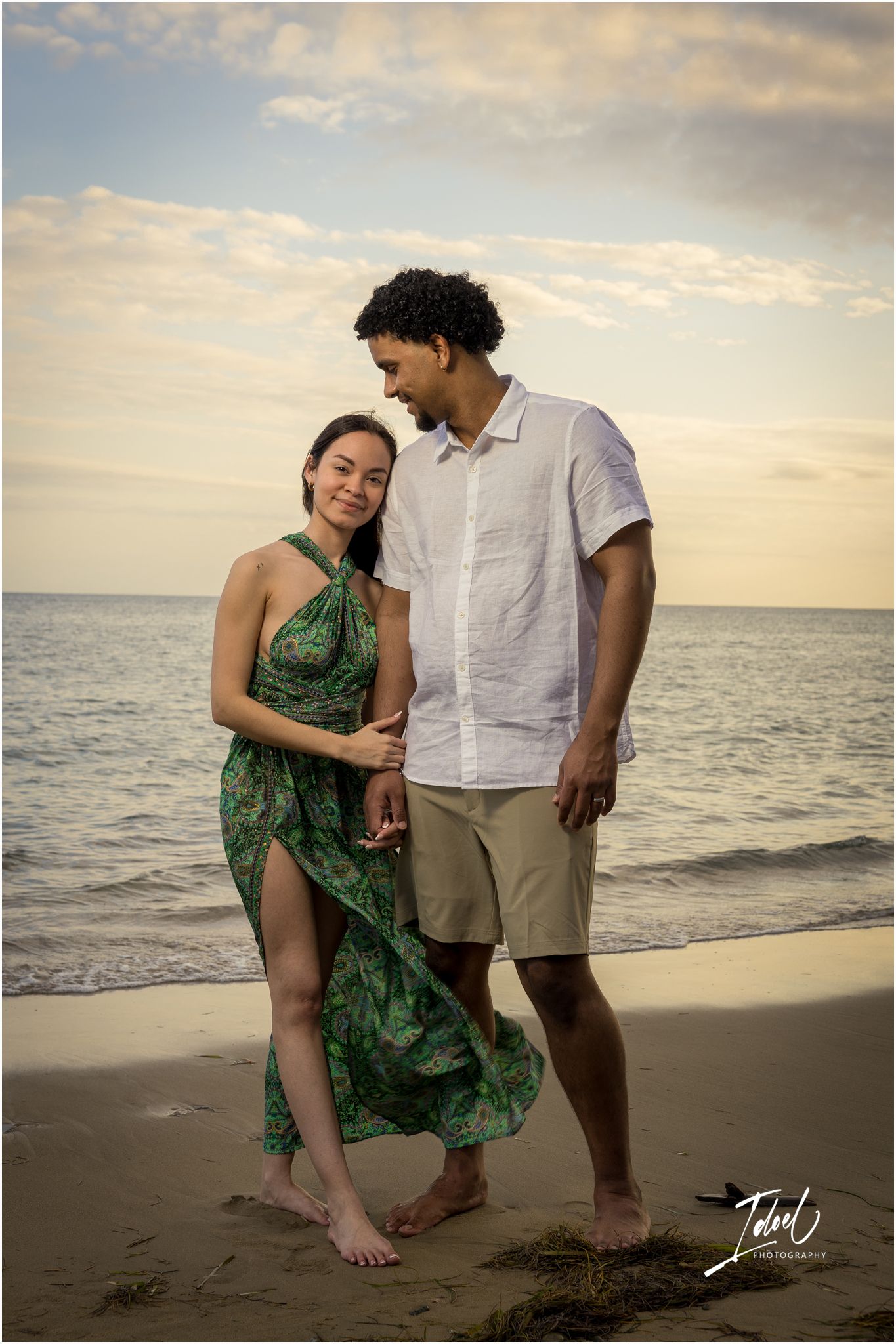 A couple shares an intimate moment at sunset on the beach, with her wearing a flowing green dress and him in white.