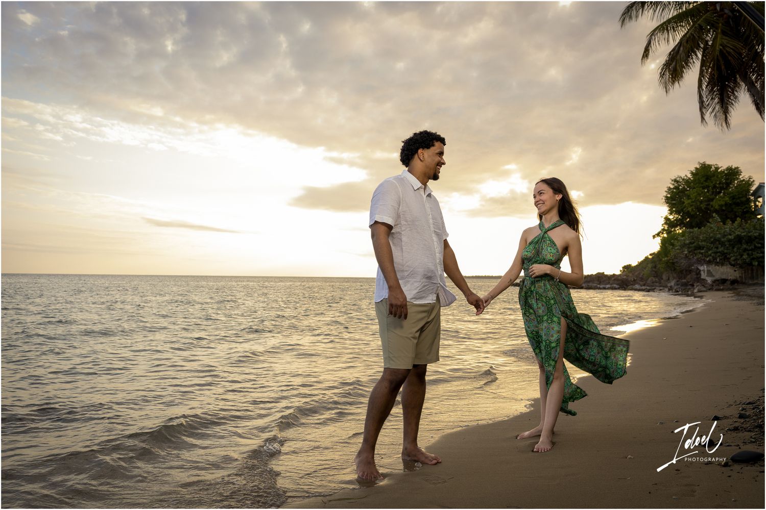 A couple holds hands while walking on a tropical beach at sunset with palm trees and ocean waves in the background.