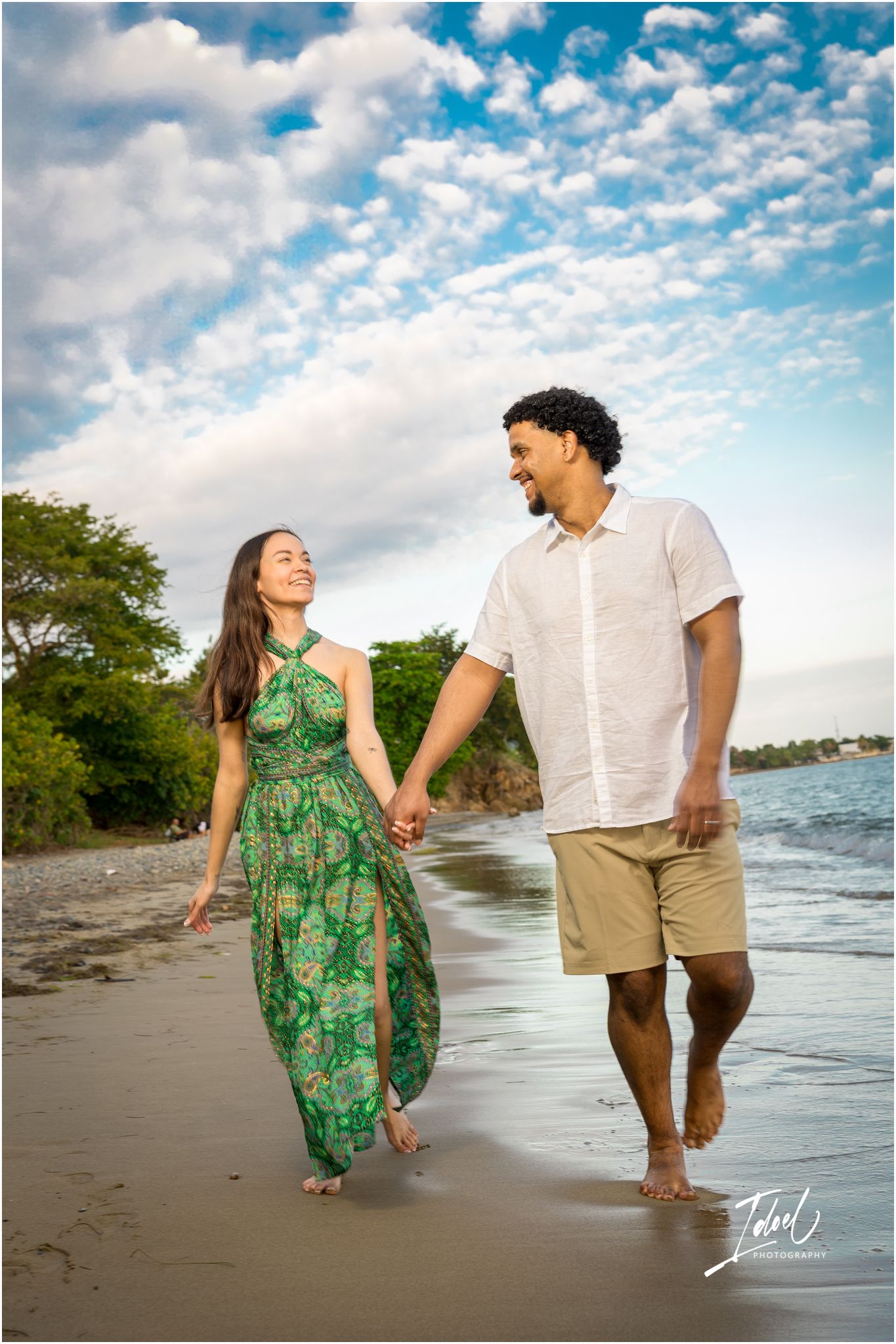 A couple walks hand in hand on a beach at sunset with puffy clouds in the bright blue sky above.
