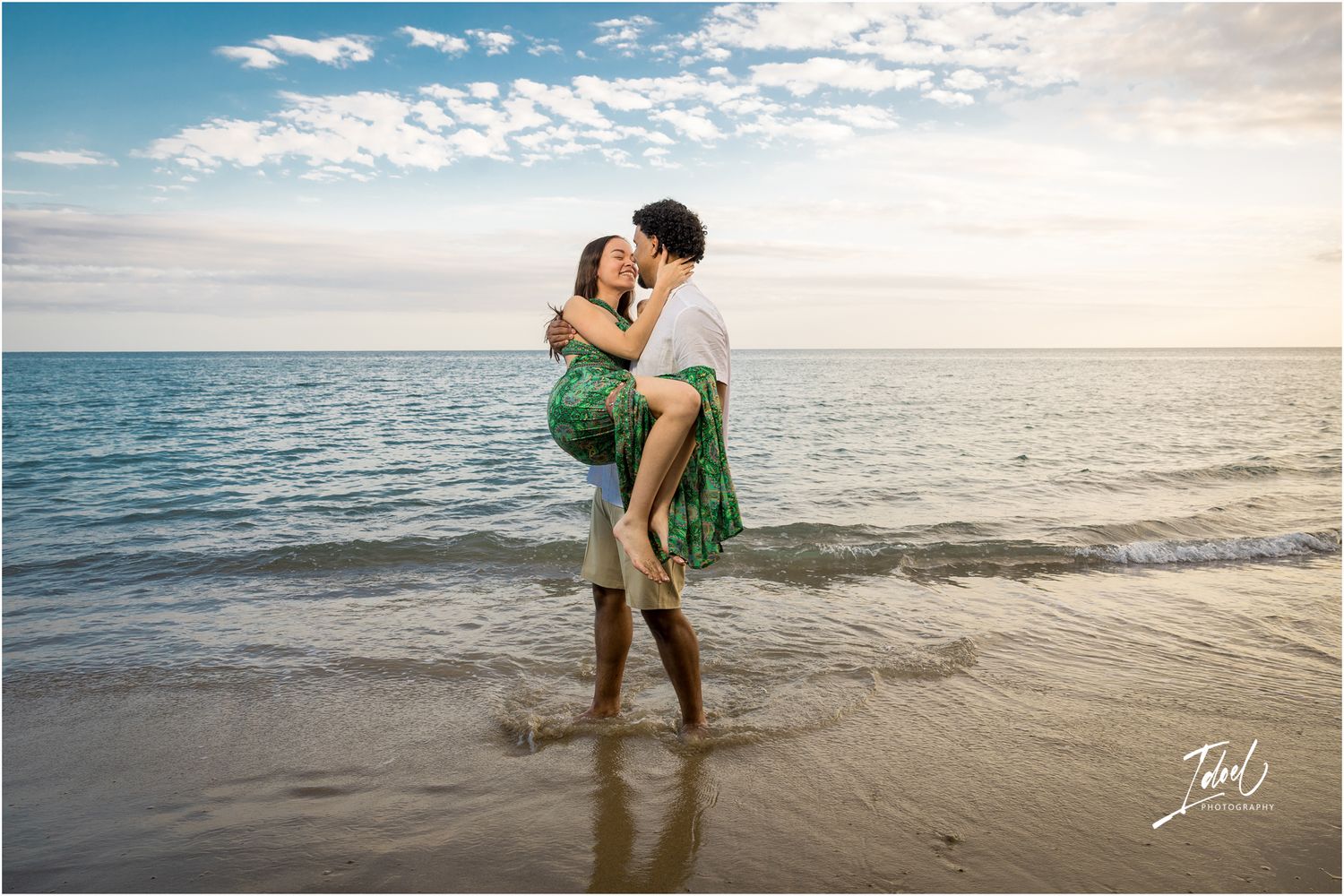 A romantic beachside embrace during sunset as waves lap at the shore and clouds dot the bright sky.