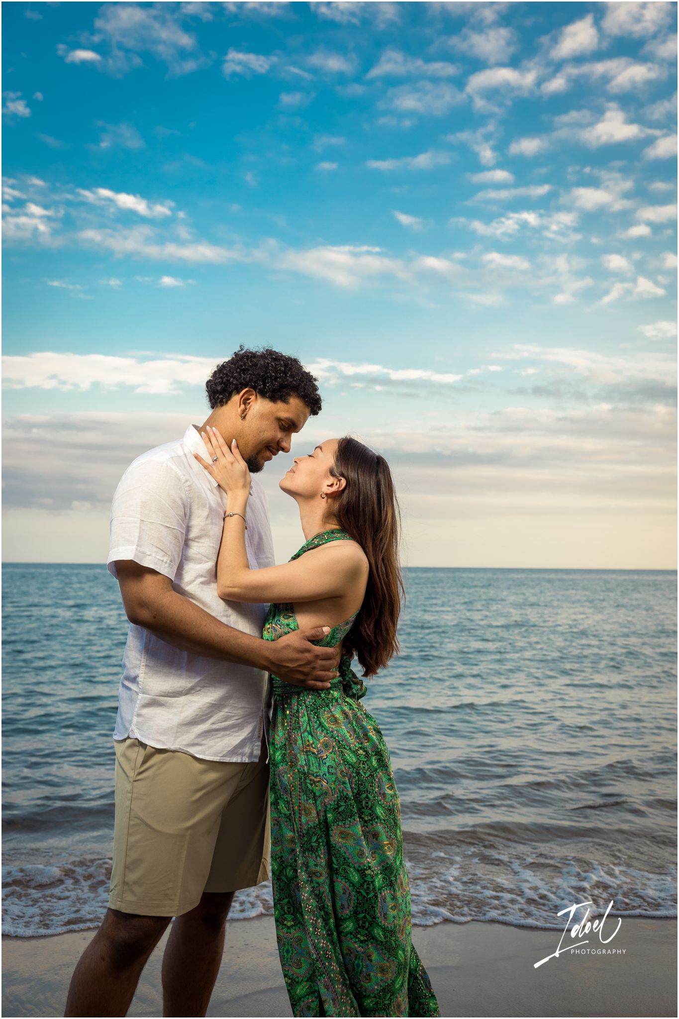 A romantic couple embraces on the beach at sunset with calm ocean waves and blue cloudy sky in the background.