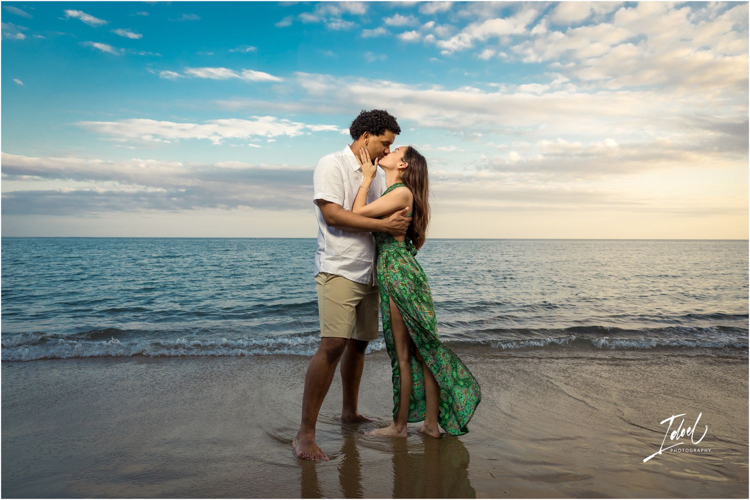 A couple embraces on a sandy beach at sunset with the ocean and cloudy sky in the background.