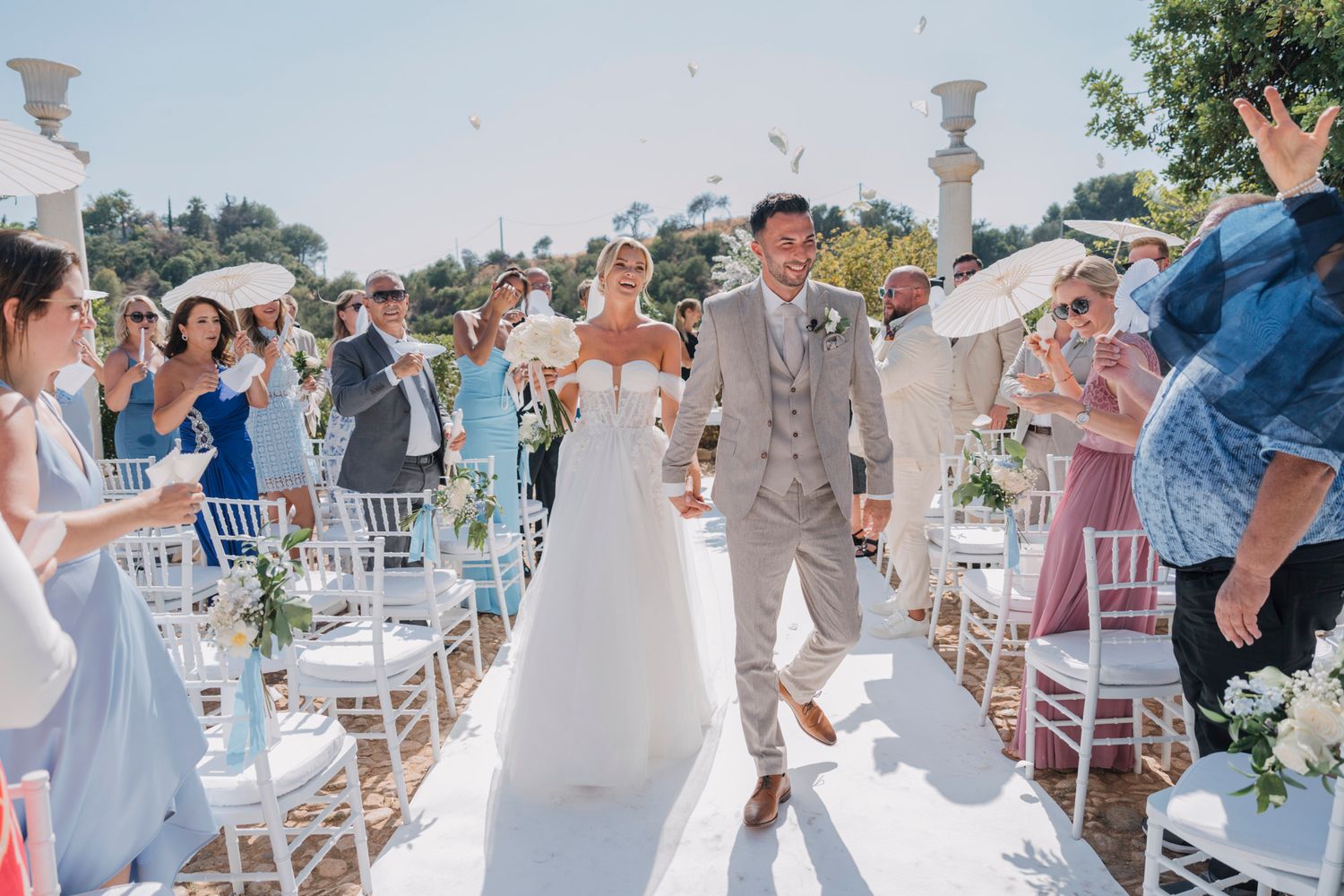 A couple walks down an outdoor wedding aisle lined with white chairs as guests smile and celebrate.