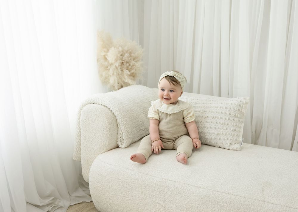 Smiling baby in cream outfit and bow headband sitting on a white boucle chaise lounge with sheer curtains.