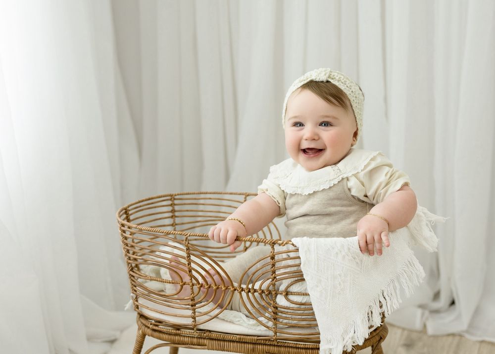 Smiling baby in cream outfit and headband sitting in a wicker bassinet against white curtain backdrop.