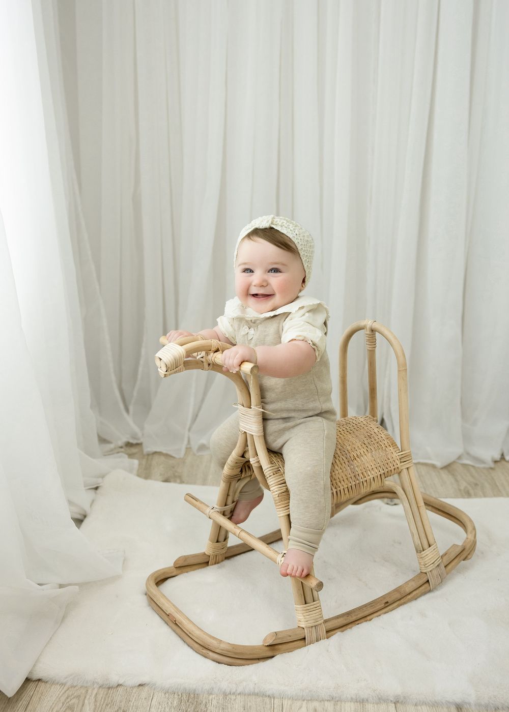 Smiling baby in beige outfit sitting on a rattan rocking horse toy on a white rug with sheer curtains.