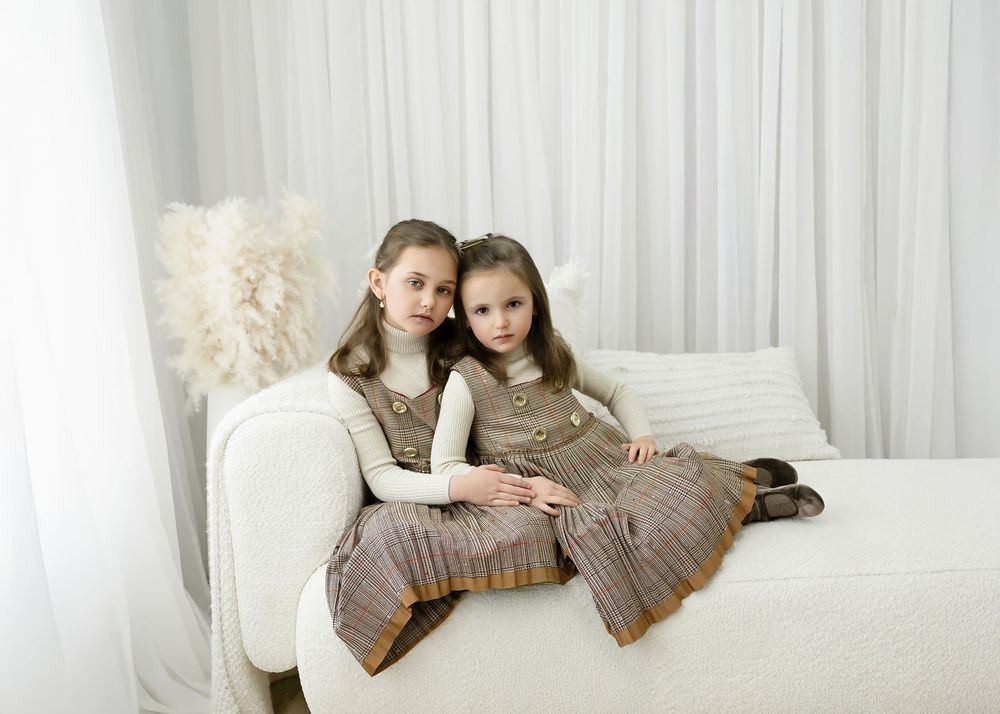 Two young girls in matching plaid dresses sit together on a white sofa in a bright, airy room.