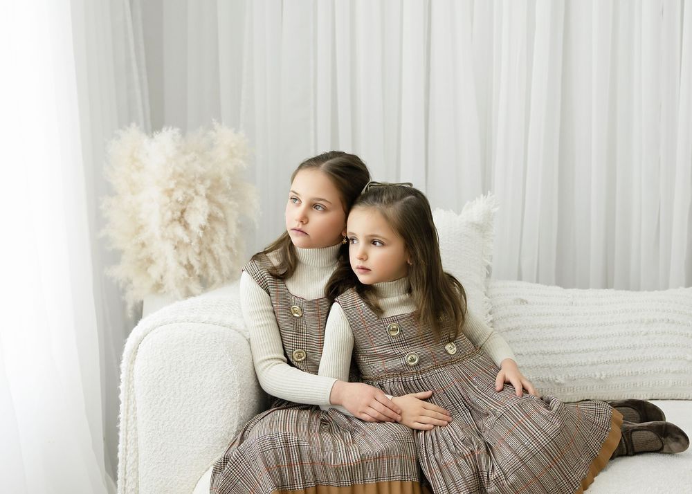 Two young girls in matching brown pinafore dresses and white turtlenecks sitting together on a white sofa.