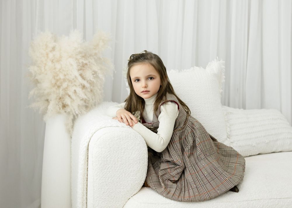 Young girl in plaid dress and white top sits gracefully on white sofa in bright, minimalist studio setting.