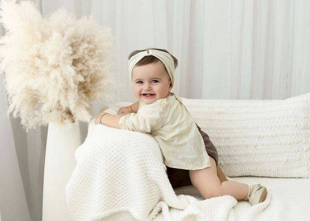 Baby wearing white knit outfit and headband lies on soft cream bedding while smiling at camera.