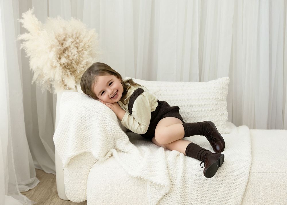 A child in a black and white outfit lies comfortably on white cushions next to a decorative pampas grass plume.