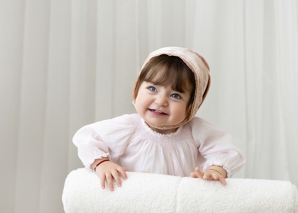 A baby in a white dress and bonnet sits and crawls in a bright studio setting across multiple poses.