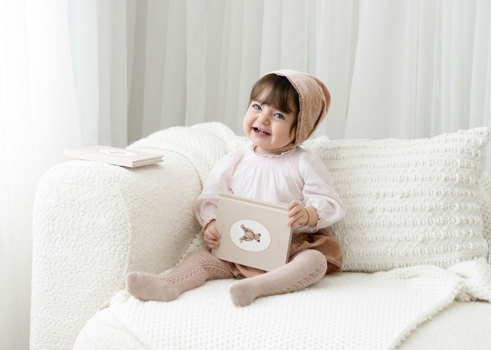 A small child in pink and beige clothing sits on a white couch with a decorative pillow.