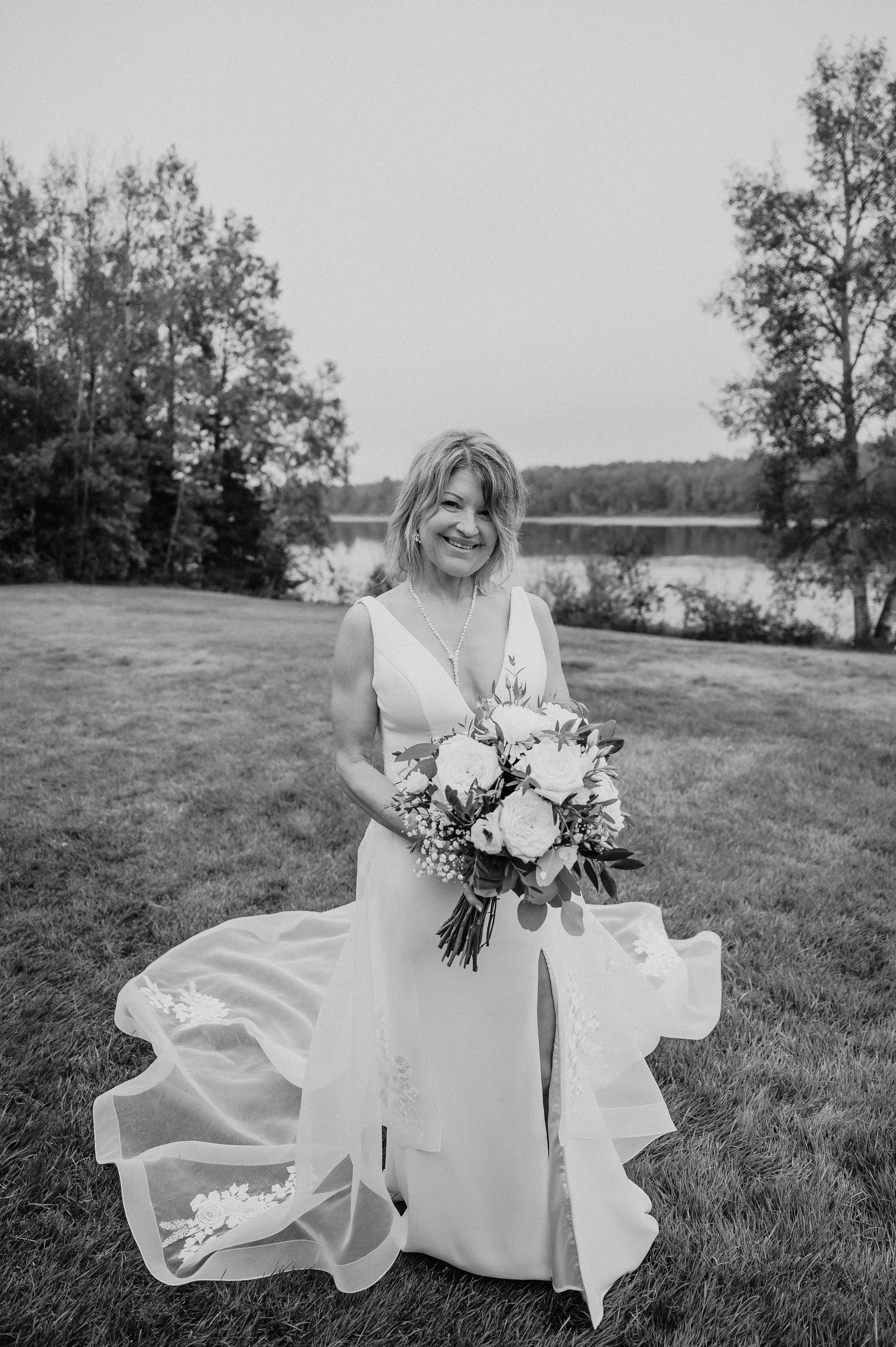 Bride with bouquet poses in flowing white dress by lakeside landscape.