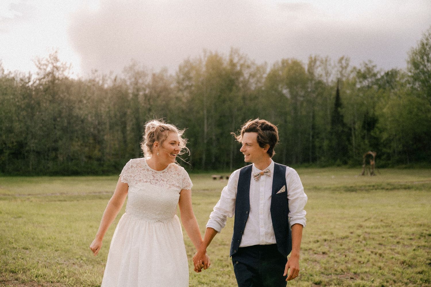 A couple holds hands while walking through a sunlit field in wedding attire.