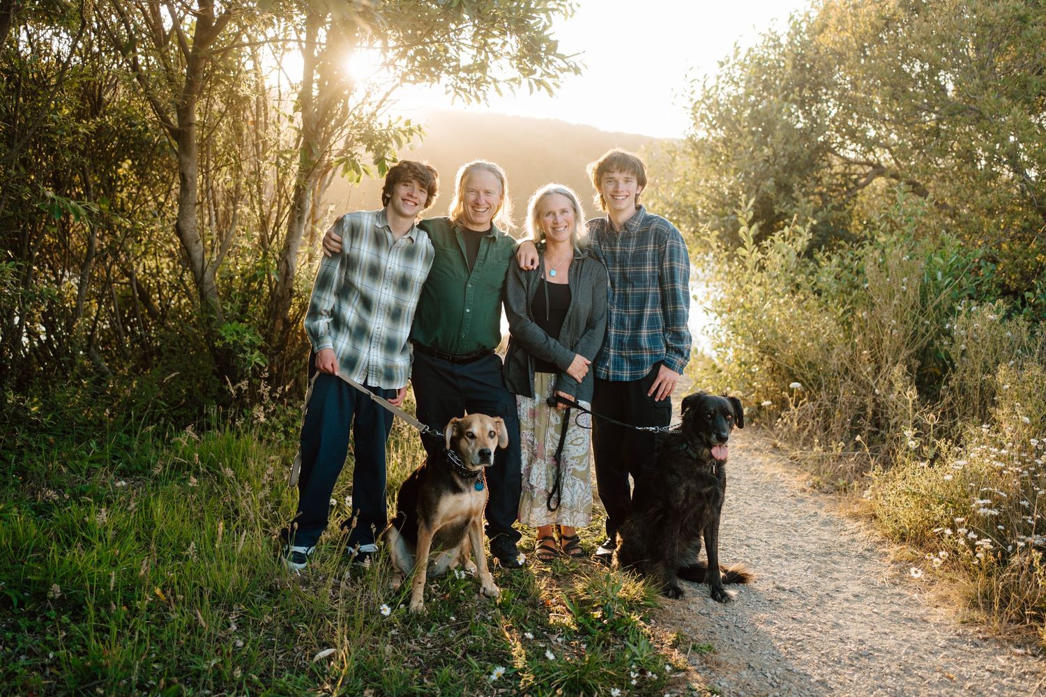 Family of four posing with two dogs on a sunlit nature trail at sunset.