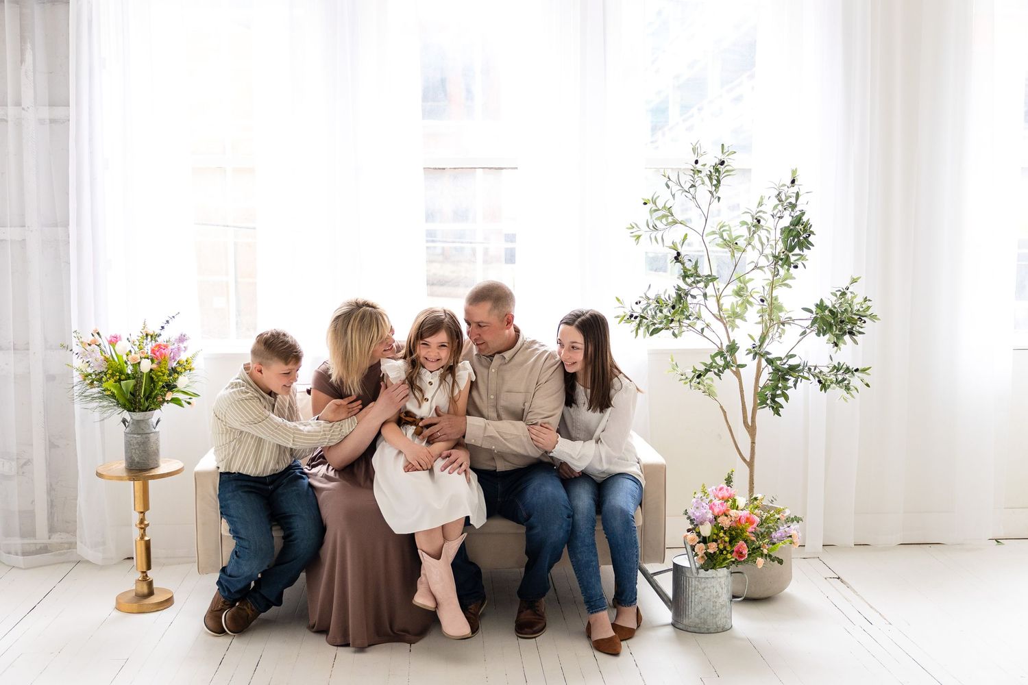 Family sitting together on white couch in bright sunlit room with flowers and indoor plant decorations.