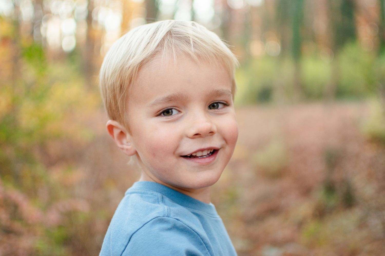 A young child in a blue shirt smiles while playing outdoors in an autumn forest setting.