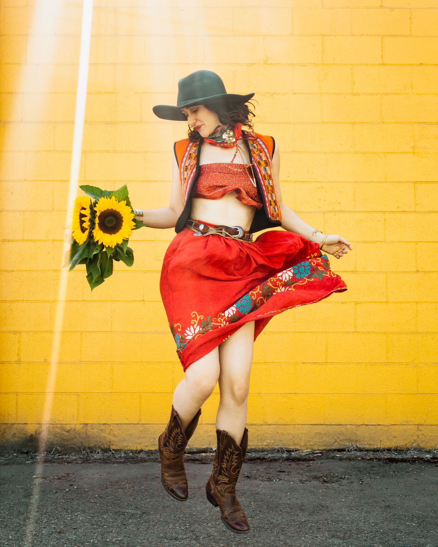 Artistic western fashion portrait against yellow wall featuring red skirt, cowboy boots, and sunflower prop in dynamic pose.