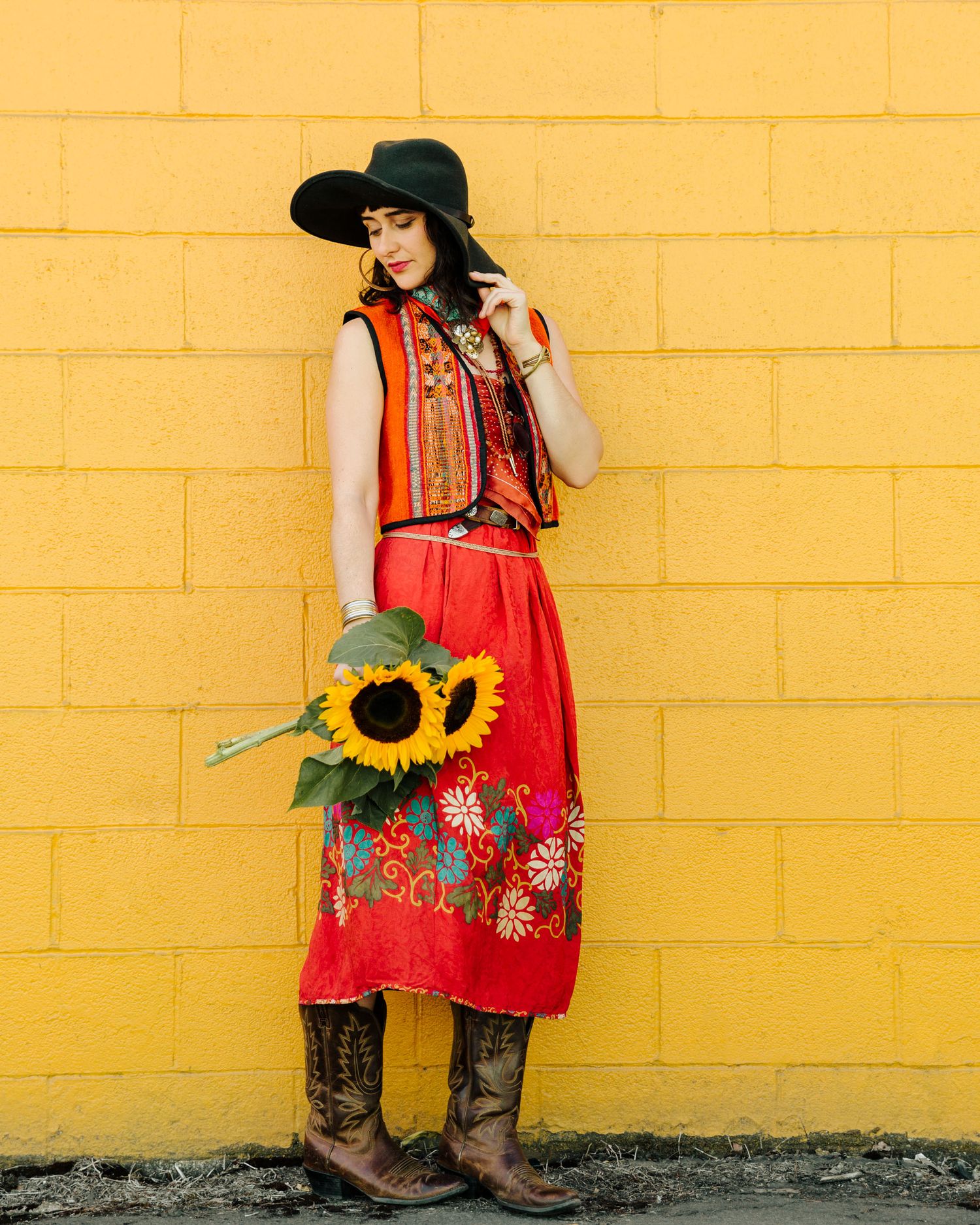 A western style outfit with red dress, vest, black hat and cowboy boots against a bright yellow wall with sunflower.