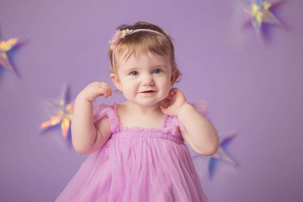 Adorable baby girl in pink tutu dress with headband smiling in front of purple backdrop with star decorations.
