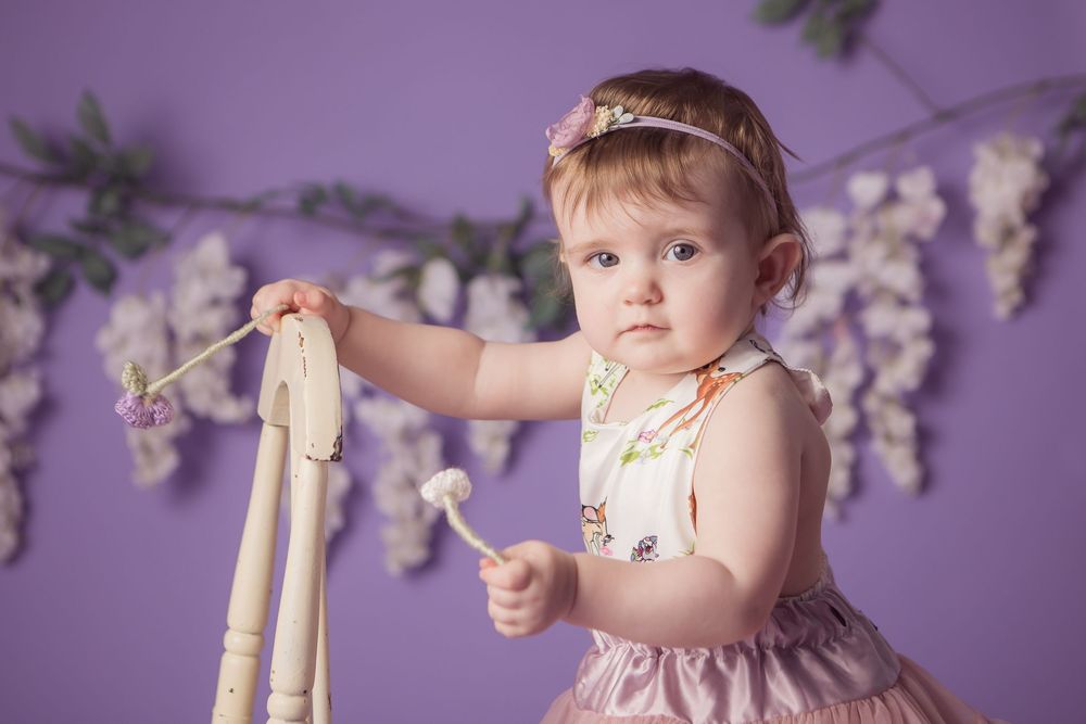 Baby girl in floral dress and headband holds white chair against purple backdrop with wisteria garland.