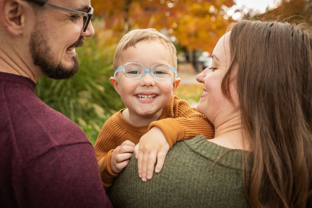 Happy toddler with blue glasses smiles between parents during autumn family portrait outdoors.
