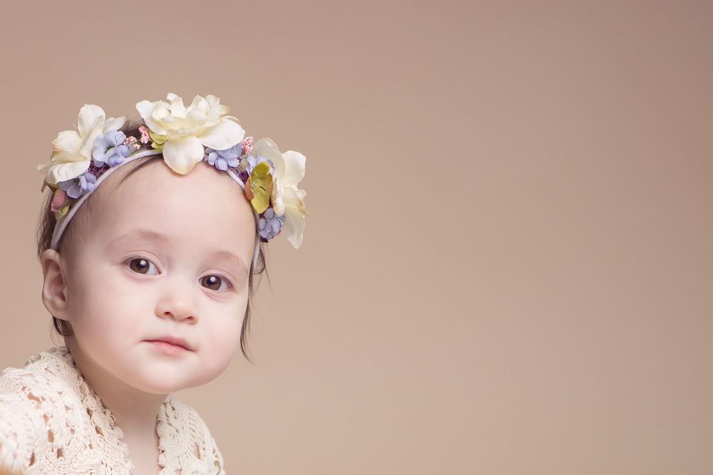 A cute baby girl wearing a floral headband with white and purple flowers, posed against a beige background.