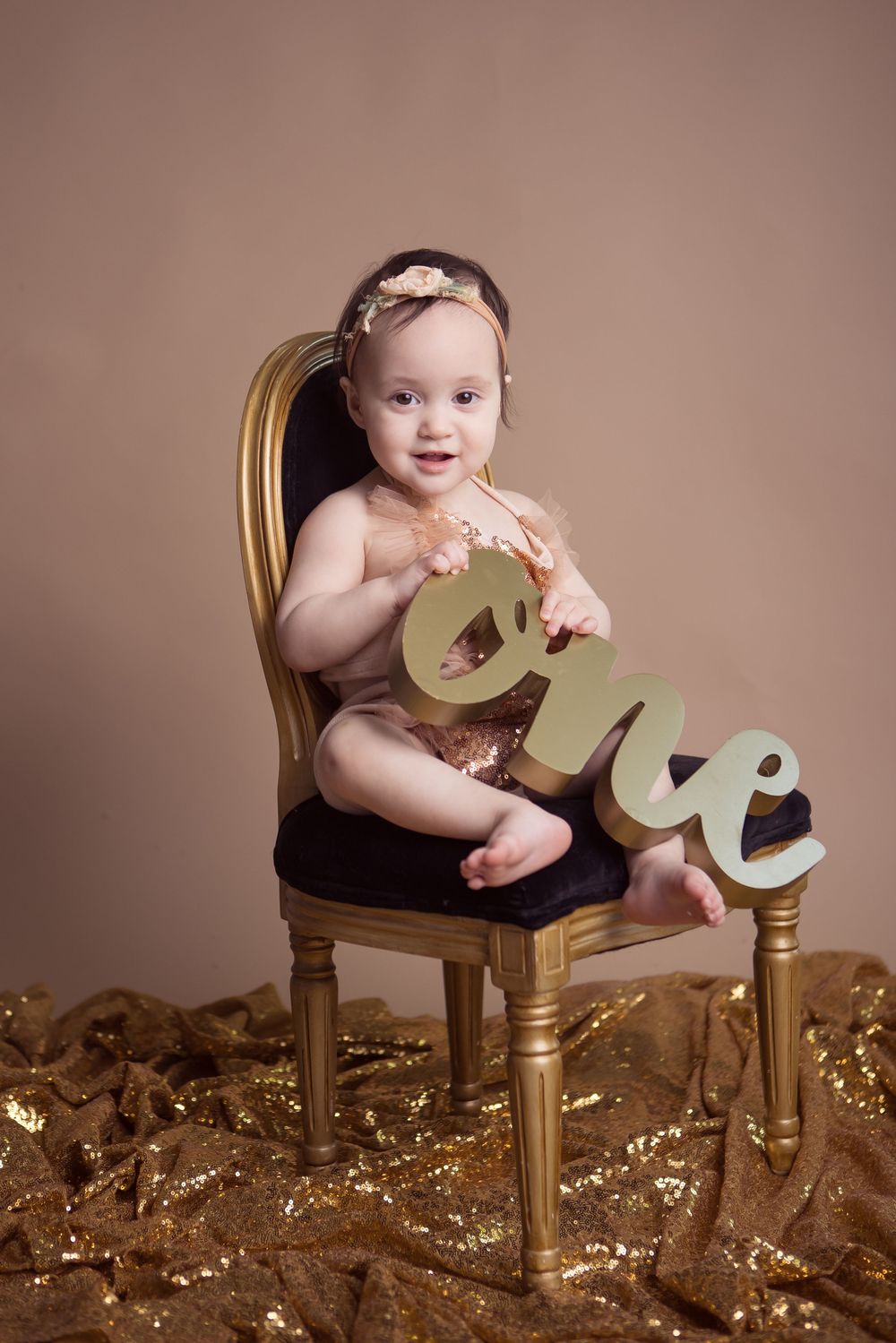 Baby girl in gold sequin outfit sits on ornate chair holding wooden “one” sign for first birthday photo shoot.