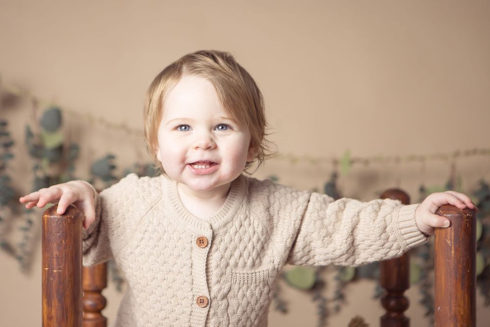 Smiling toddler in beige knit cardigan standing at wooden chair rails with eucalyptus garland backdrop.