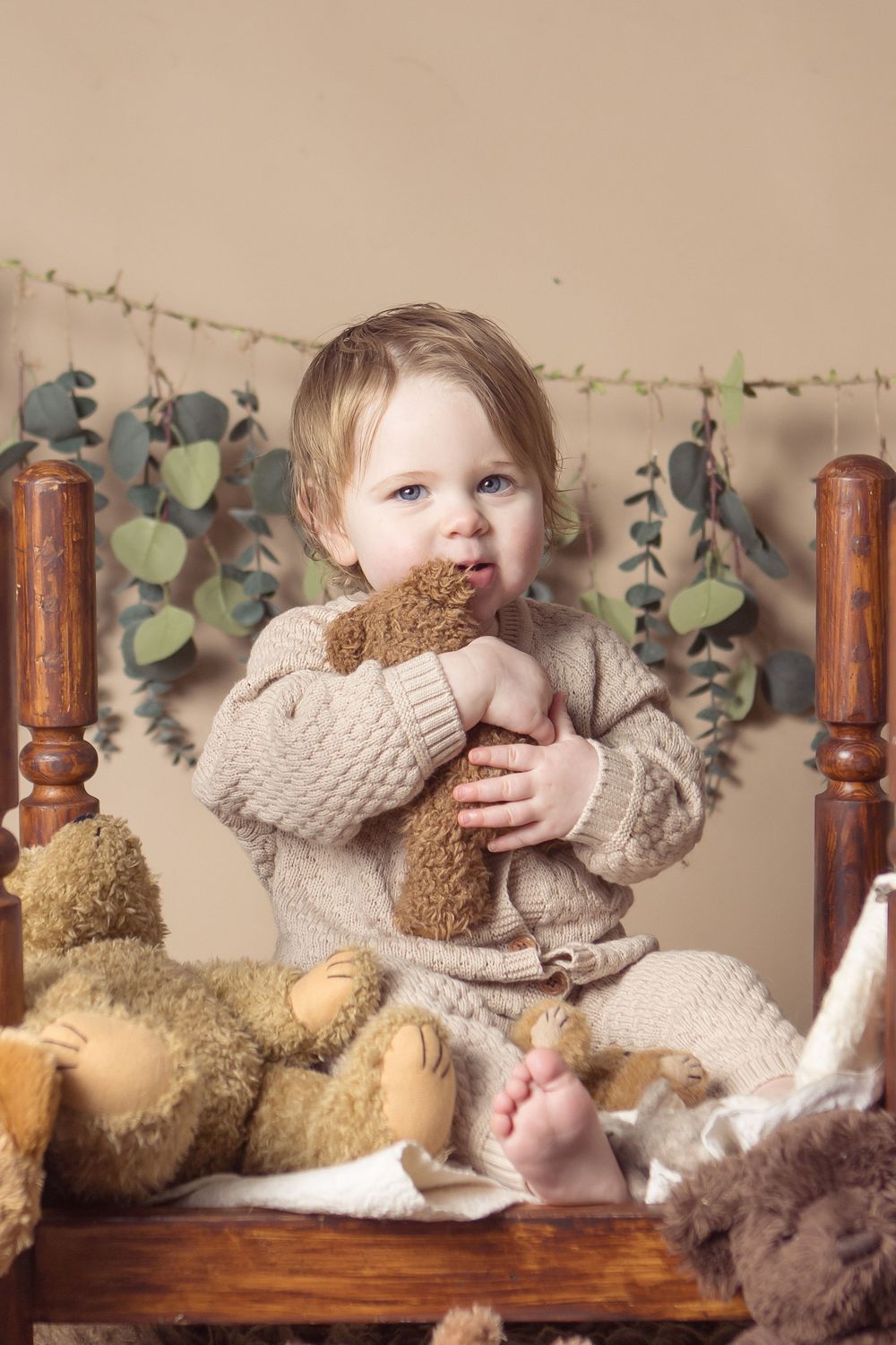 Toddler in beige knit sweater hugging a teddy bear on wooden chair with eucalyptus garland backdrop.