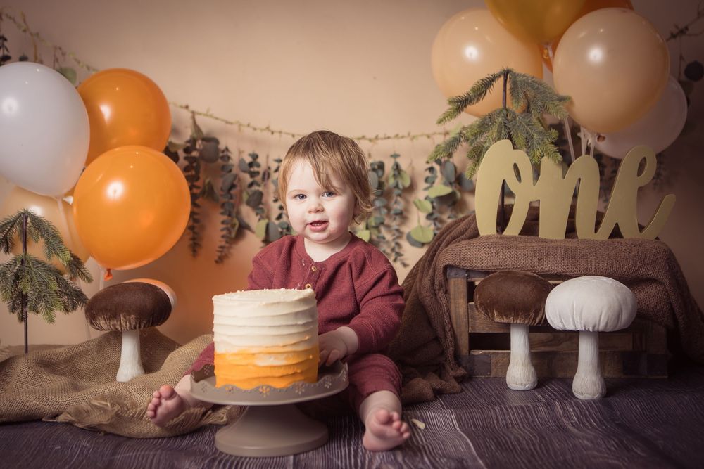 Toddler in red outfit sits before ombre birthday cake surrounded by orange balloons and woodland decor for first birthday smash.