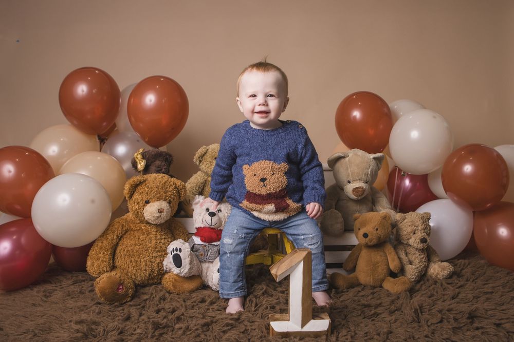 Baby boy in blue bear sweater sits smiling among teddy bears and balloons beside a number one for his first birthday.
