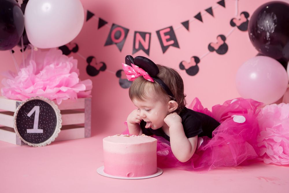 Baby girl in pink tutu eats first birthday cake at Minnie Mouse themed party with balloons and ONE banner.
