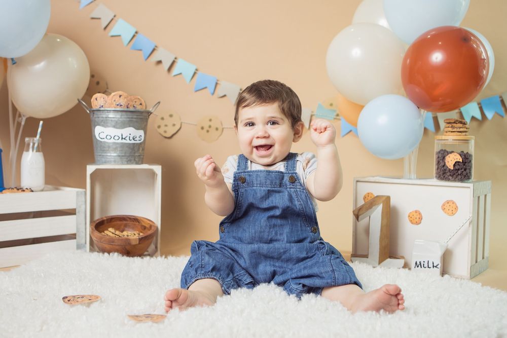 Baby in denim overalls smiling at first birthday cake smash with balloons, cookies, and milk bottle decorations.