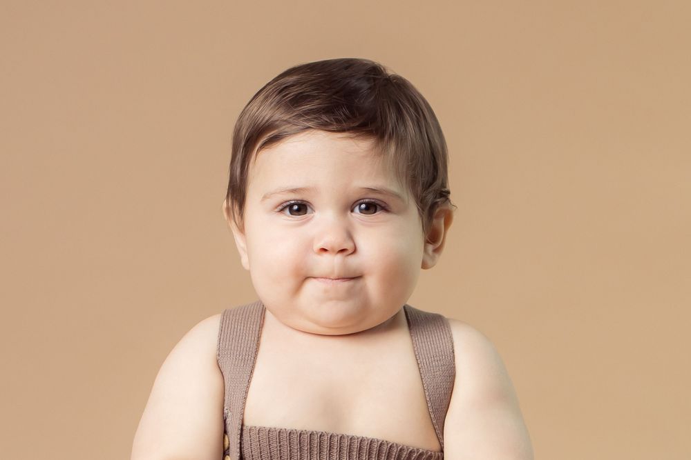 Adorable baby in brown overalls posing against a beige background with a cute expression.