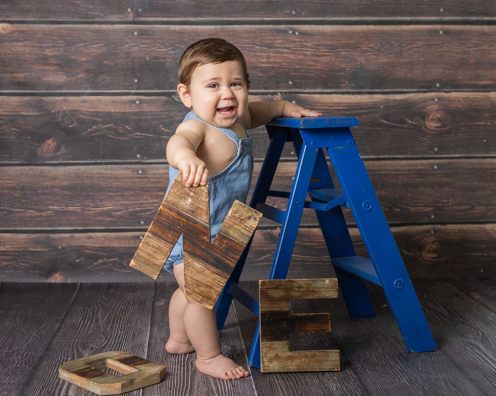 Laughing baby in denim overalls sitting beside a blue ladder with wooden blocks on a rustic wood floor.
