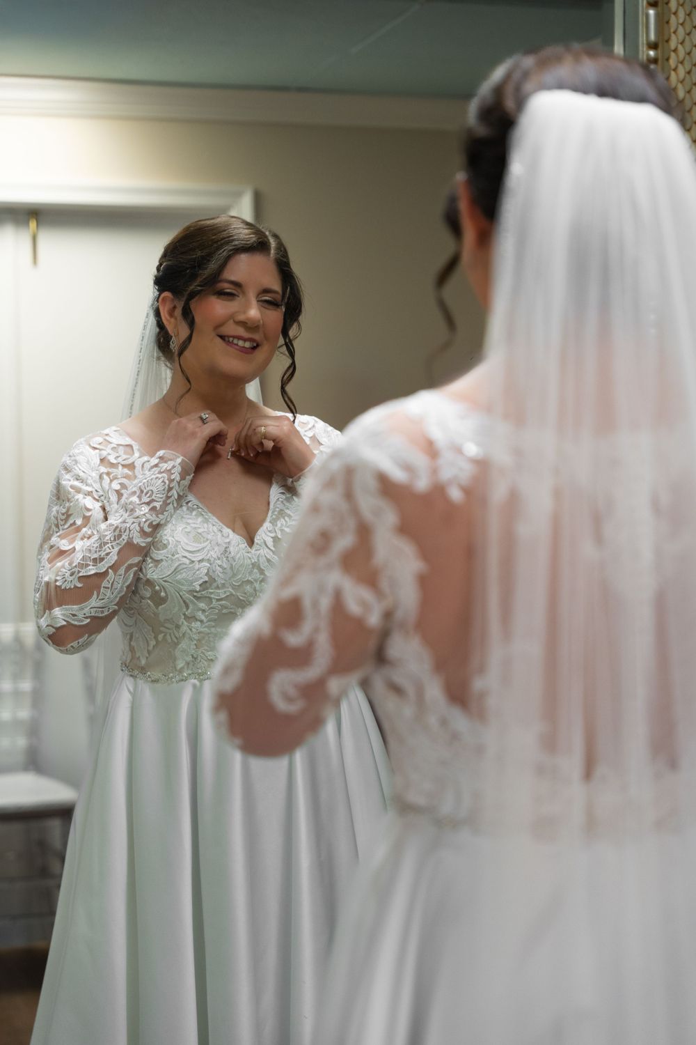 Bride adjusting her necklace in front of a mirror, smiling softly in her lace wedding dress, bridal preparation moment.