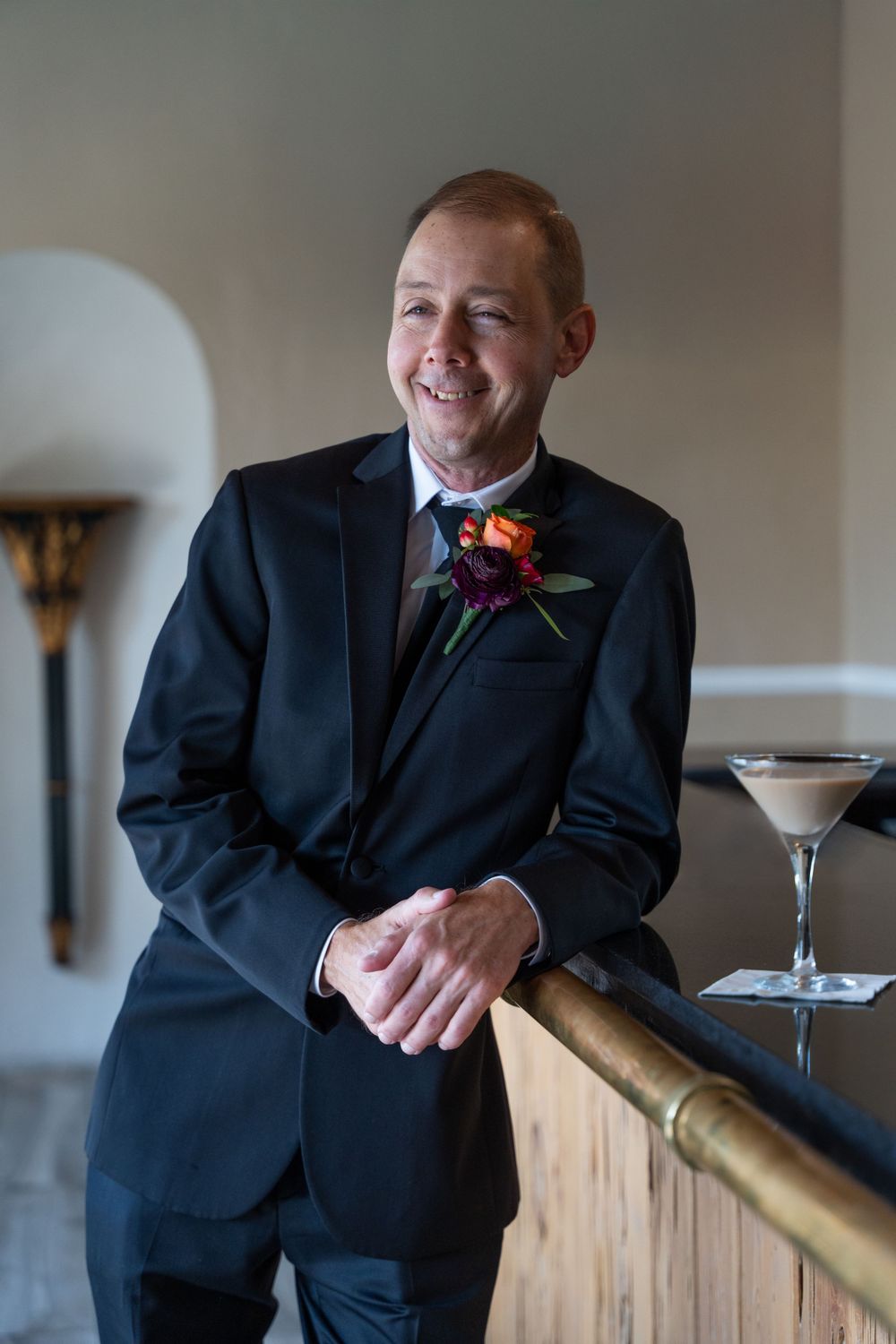 Groom in a black suit with a vibrant boutonniere, smiling confidently by the bar with a cocktail, wedding day portrait.