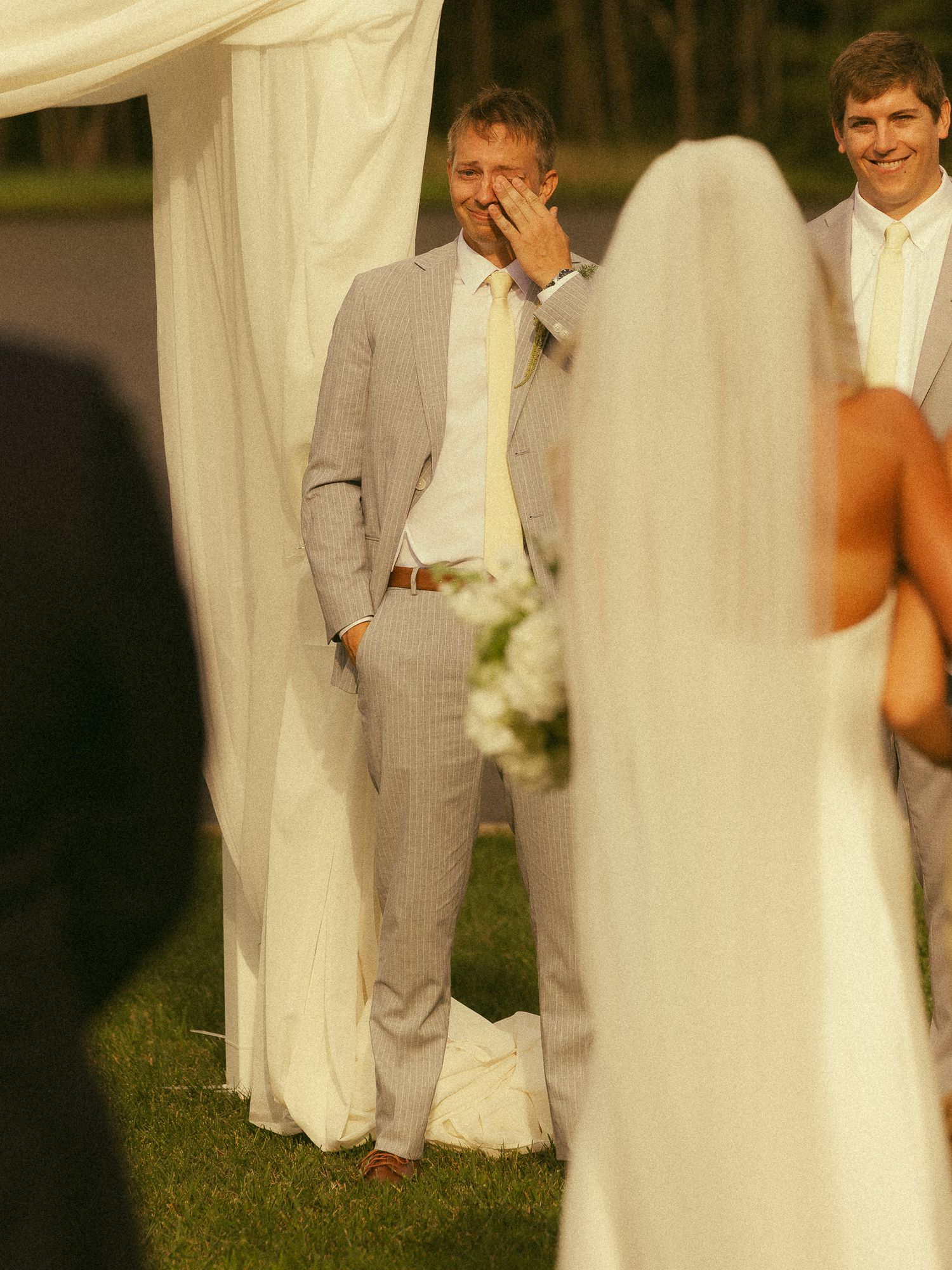 Emotional groom wiping tears as bride approaches in outdoor wedding ceremony with white draping.