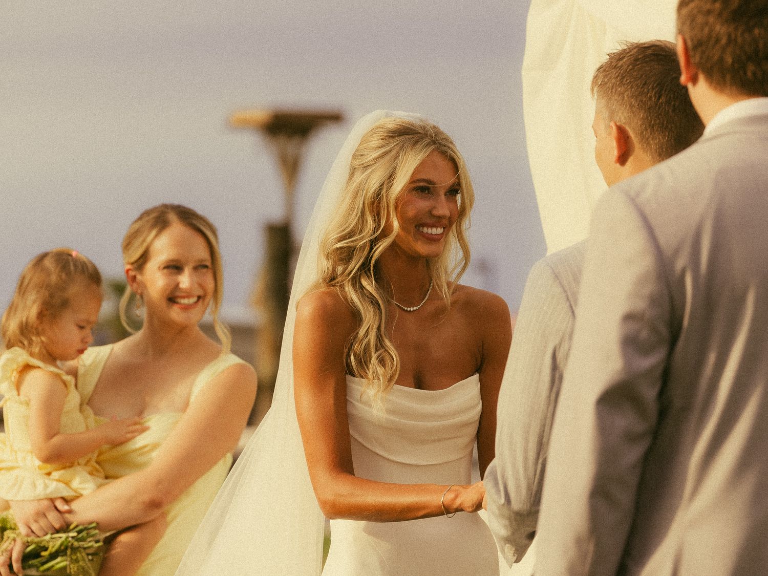 Smiling bride in white strapless gown holds hands with groom at outdoor wedding ceremony with bridesmaid and flower girl.