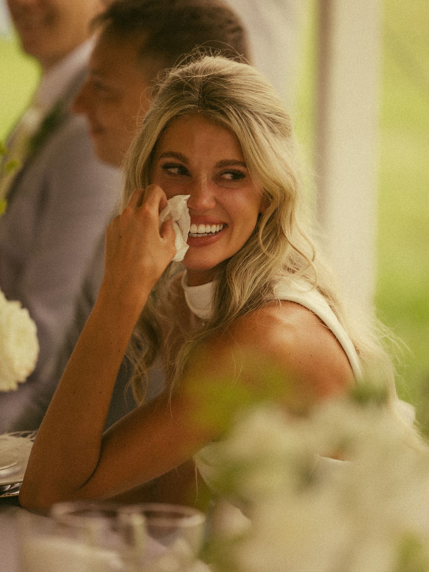 Blonde woman smiling and wiping tears of joy at an outdoor wedding reception, seated at a floral decorated table.