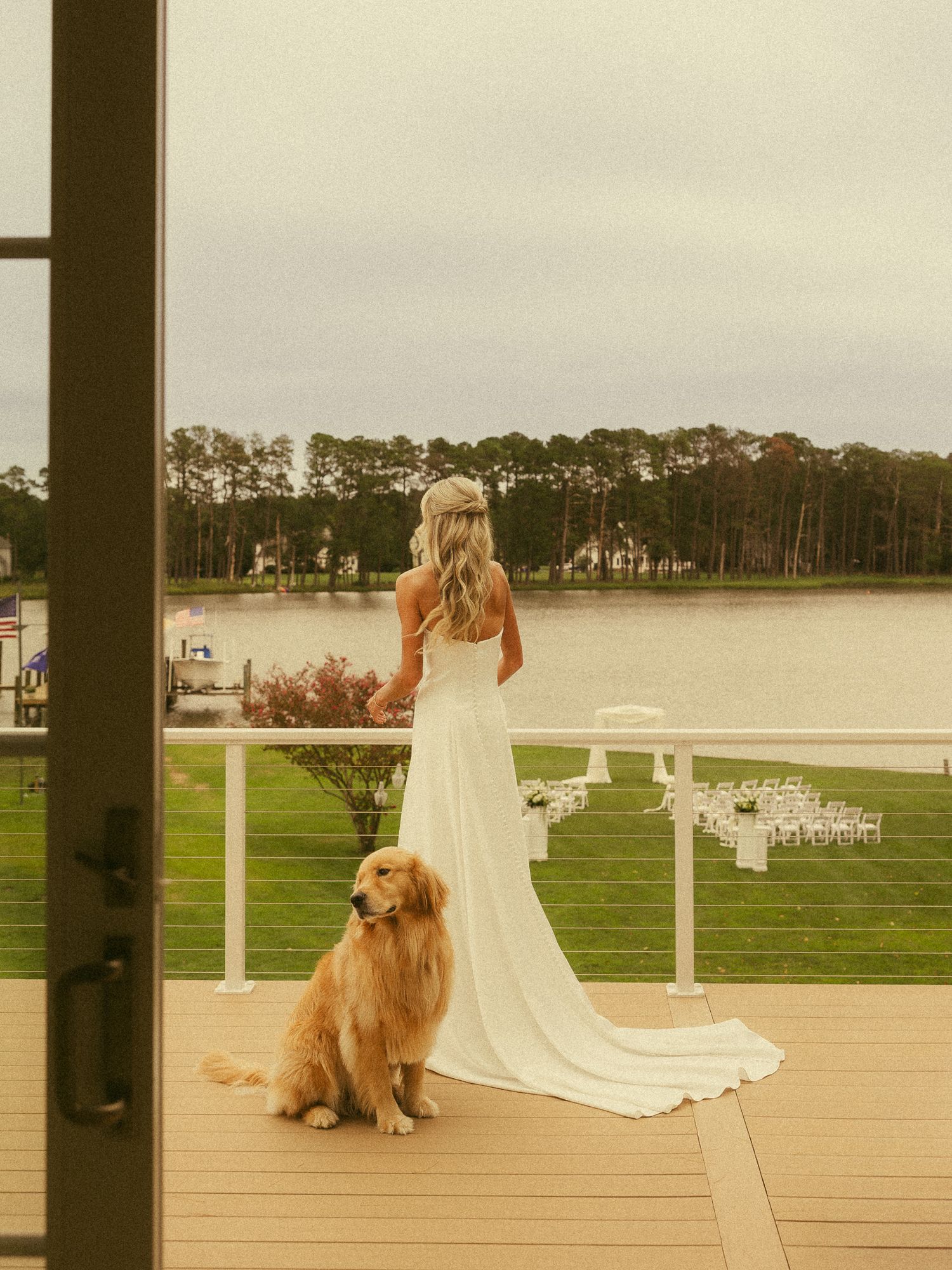 Bride in white gown stands on waterfront deck with golden retriever, overlooking outdoor wedding ceremony setup.
