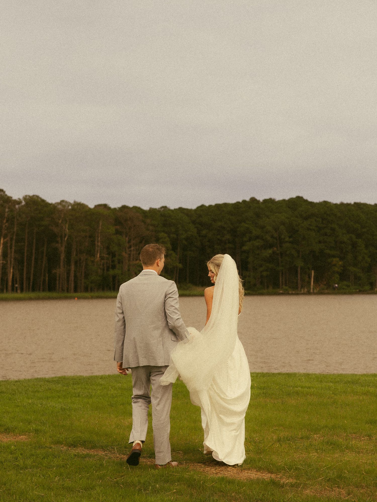 A bride and groom walk hand in hand toward a serene lakeside, surrounded by lush green trees on their wedding day.