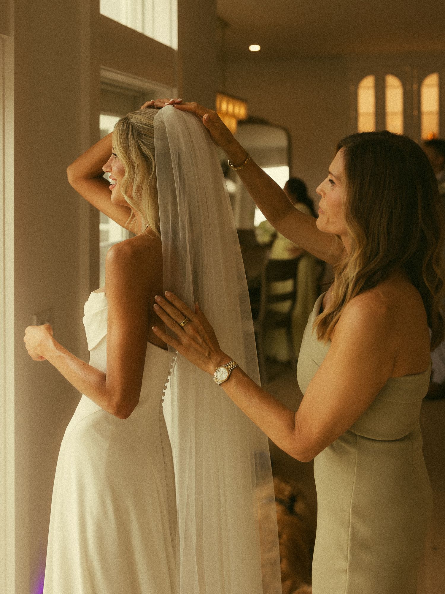 A bridesmaid helps a smiling bride adjust her white veil while getting ready indoors in warm golden light.
